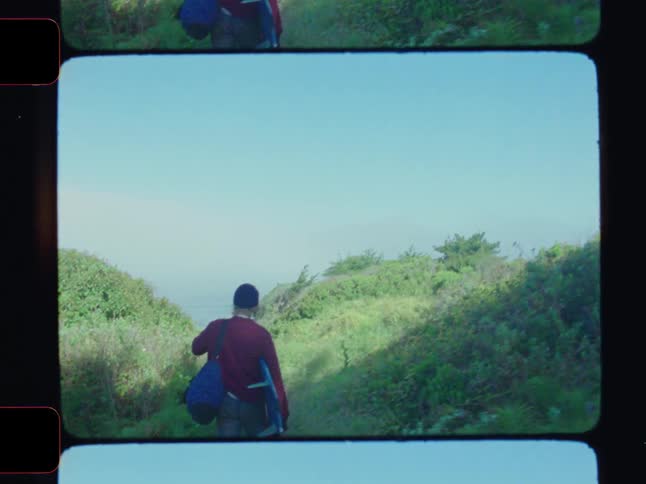 A blonde man carries a surfboard along a dirt path toward the ocean on a sunny summer day
