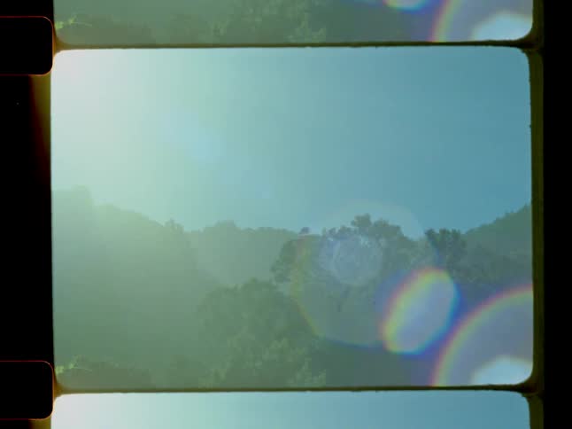 A hazy sunlit landscape view with green hills and a prominent lens flare is displayed within a film frame