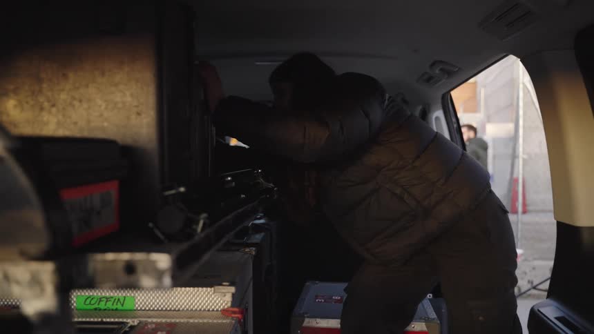 Woman stands in a van examining equipment while others stand next to a building nearby