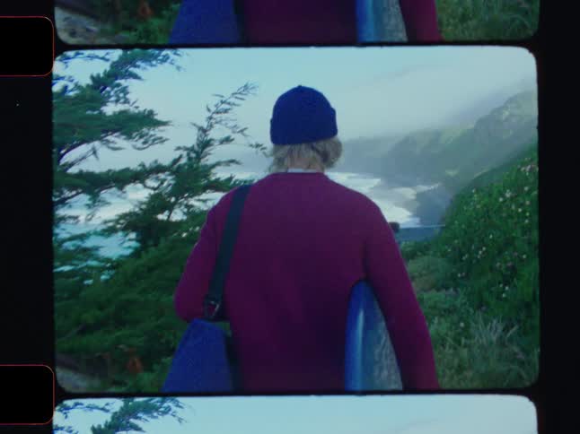Man in a cap carrying a surfboard along a trail towards the sea