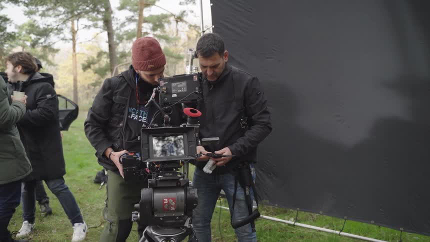 Two camera operators adjust a professional film camera on a tripod during an outdoor shoot with other crew members nearby and a large black backdrop