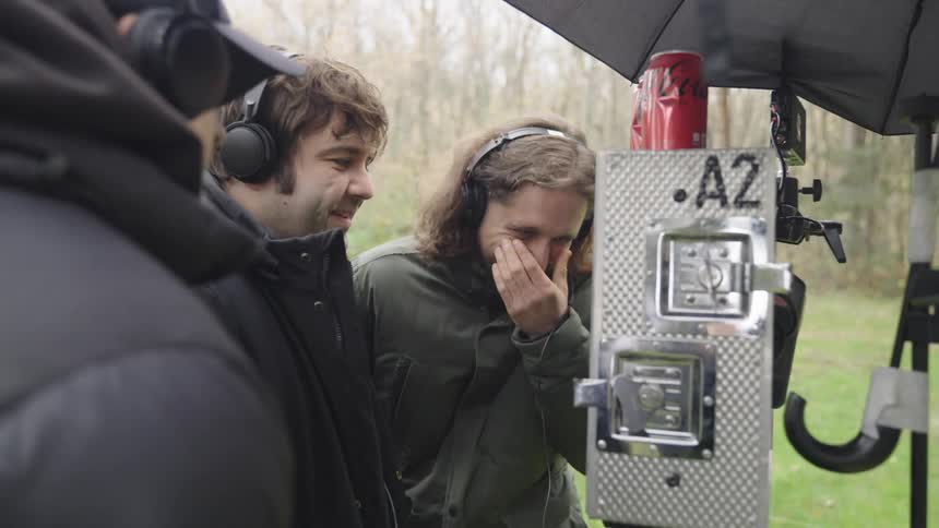 Three men laughing as they stand on an outdoor recording set in front of an instrument
