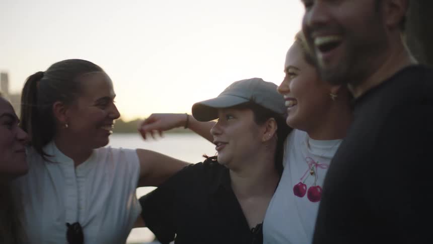 A group of young friends laughs claps and hugs joyfully outdoors near water with a city skyline in the background during sunset