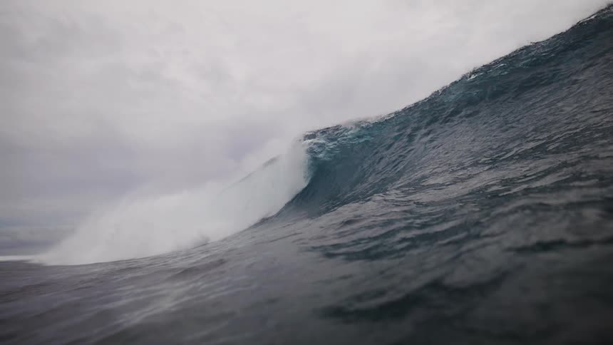 A powerful dark blue wave possibly curls and breaks under a cloudy sky