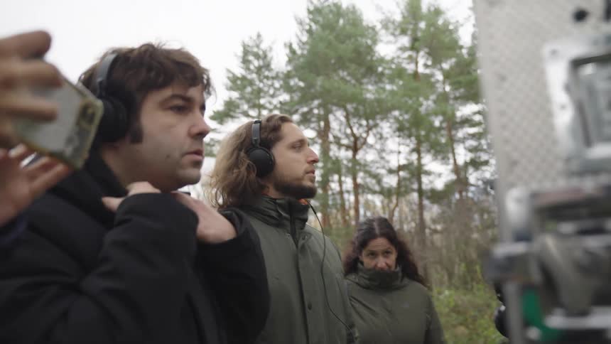 Two men put on headphones to listen joined by a woman standing near audio equipment on an outdoor film set in the woods