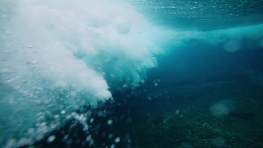 An underwater view of a turbulent mass of white foam and bubbles from a breaking wave crashing into the clear blue water with a glimpse of the dark seabed below