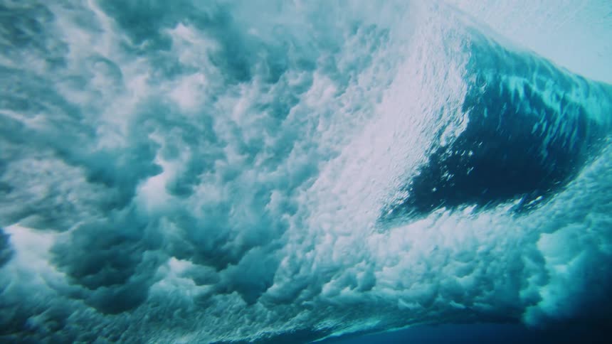 An underwater view captures the chaotic energy of a breaking wave filled with foamy light blue water against the darker blue depths below