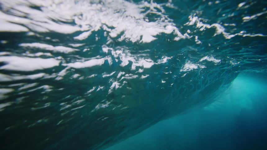 An underwater view looks up at the turbulent surface of the water where a wave is breaking and creating foamy textured patterns against the clear blue water and the darker depths below