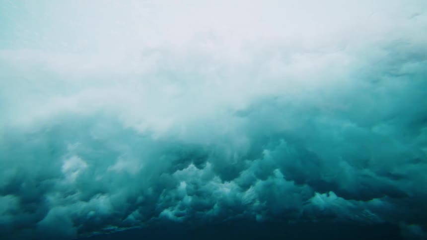 An underwater view shows the surface of the water above where sunlight penetrates and illuminates the clear turquoise water before it fades into the deep dark blue below