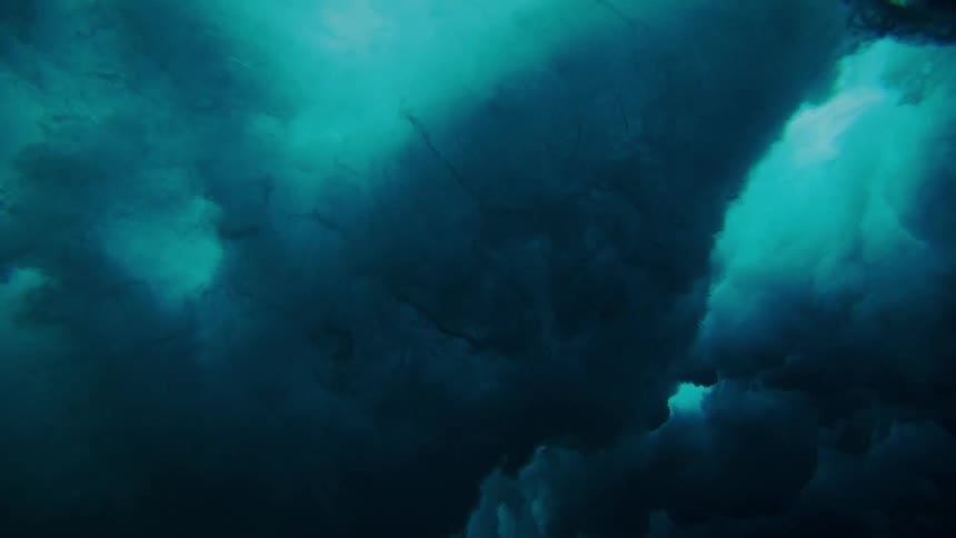 An underwater view shows the turbulent foamy underside of a breaking wave as it crashes into the deep blue ocean above a dark seabed