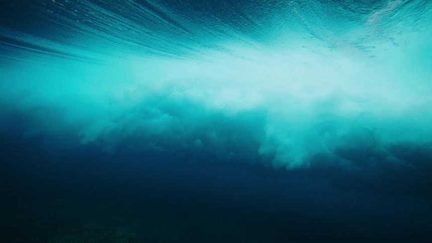 An underwater view captures the dynamic interplay of light and shadow as waves break above creating streaks of bright turquoise against the deep dark blue of the water below
