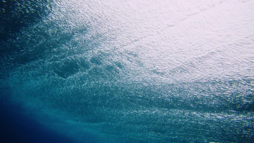 An underwater perspective looks up at the surface of the water where sunlight creates bright shimmering patterns against the deep blue below