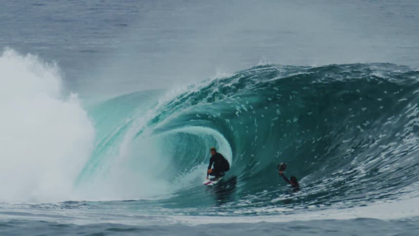 A surfer in a black wetsuit skillfully rides inside the barrel of a large turquoise wave as it breaks and churns white water under a cloudy sky