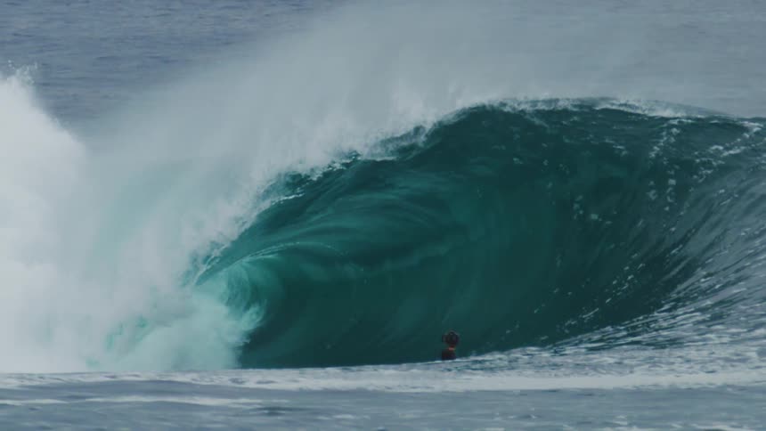 A surfer in a black wetsuit stands on a jet ski in the ocean looking at a massive wave that is starting to break and create a large spray of white water under an overcast sky