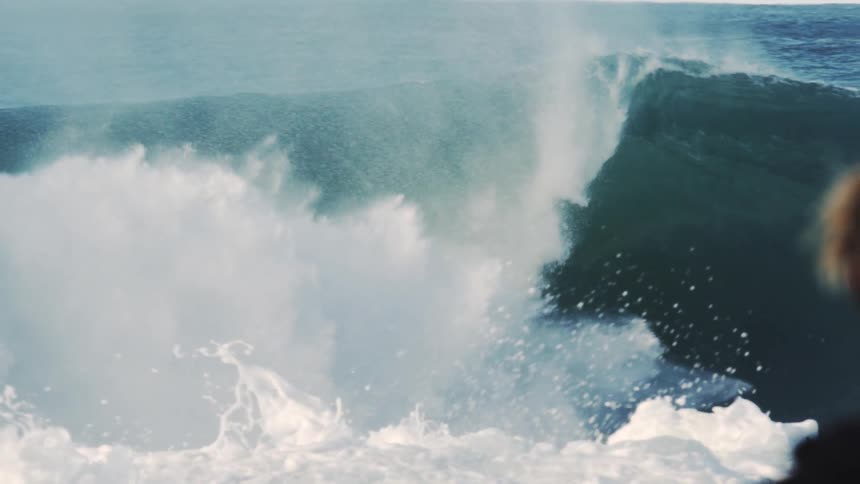 Man stands watching surfers as a large breaking wave sprays water into the air