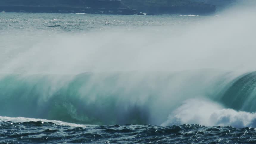 A large powerful dark teal wave crests and begins to break creating a foamy white spray against the backdrop of the deep blue ocean and a distant coastline