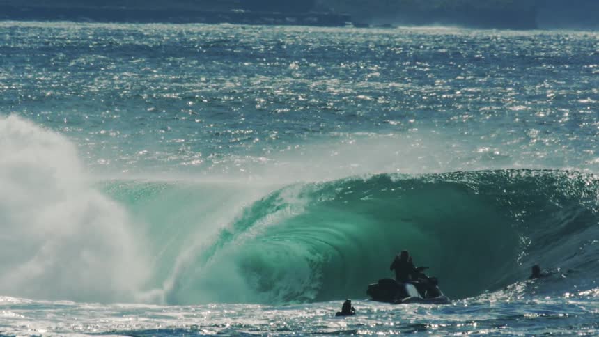 Person on jetski following surfers in turquoise blue sea with tubular waves