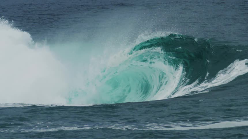 Captured in slow motion a massive turquoise ocean wave curls and crashes powerfully engulfing a lone surfer near its base