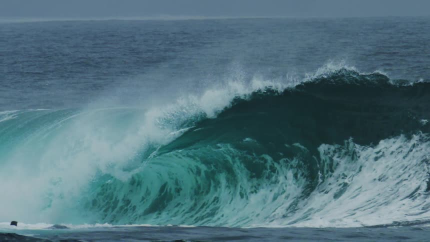 A massive turquoise ocean wave curls powerfully creating a tube and sending spray into the air as a lone surfer paddles nearby before it crashes down