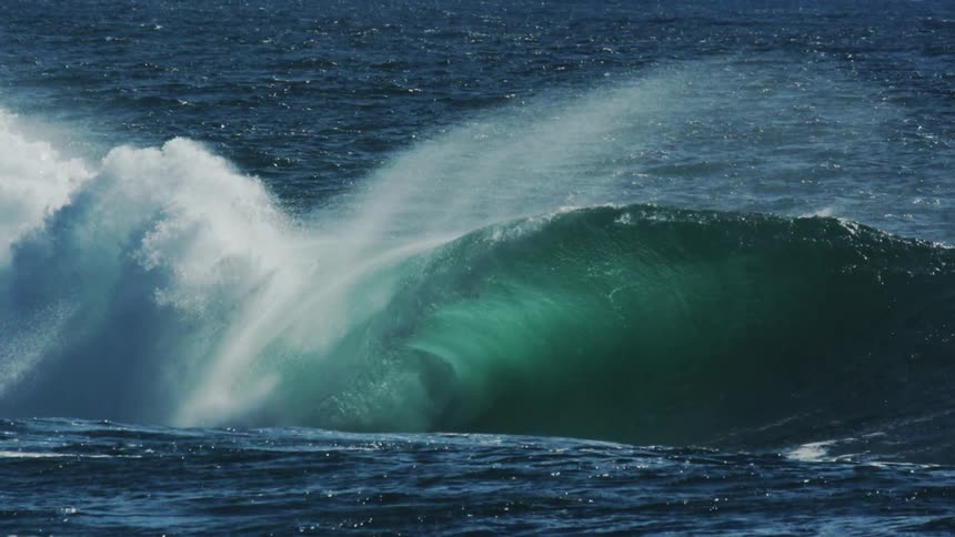 A powerful emerald green wave crashes and sprays white water against the deep blue surface of the ocean under a bright sky
