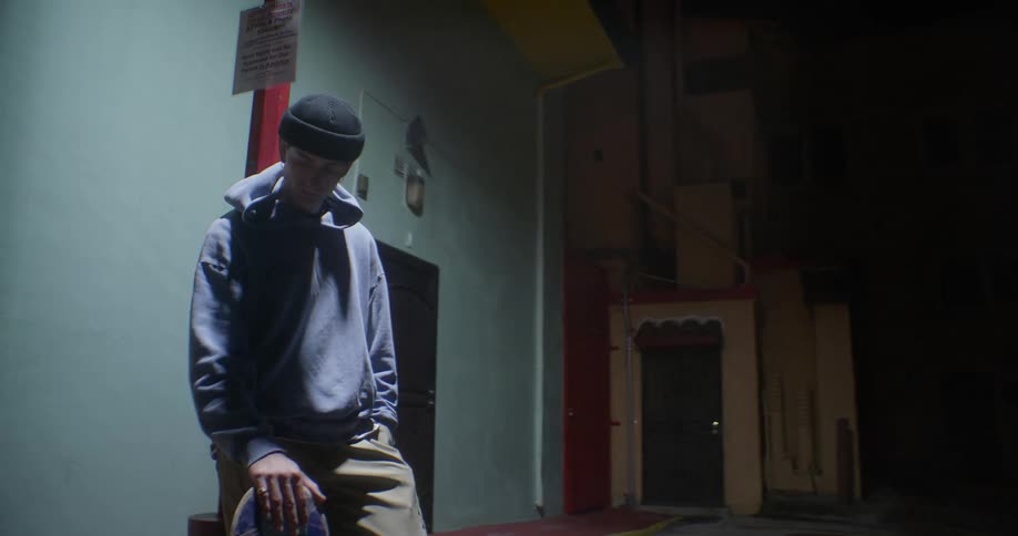 Young man in an alley at night holding a skateboard and leaning against a lamppost
