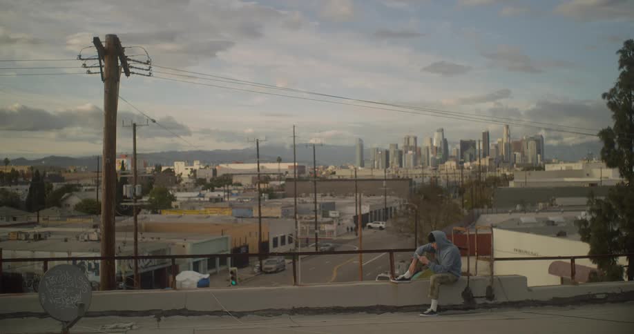 Youth sits on the roof of a building looking at his smartphone on a cloudy day with the city skyline in the background