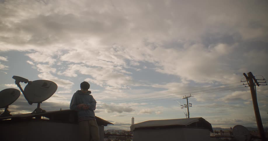 A person stands on an urban rooftop looking down at a smart phone framed against a cloudy sky with satellite dishes and power lines nearby