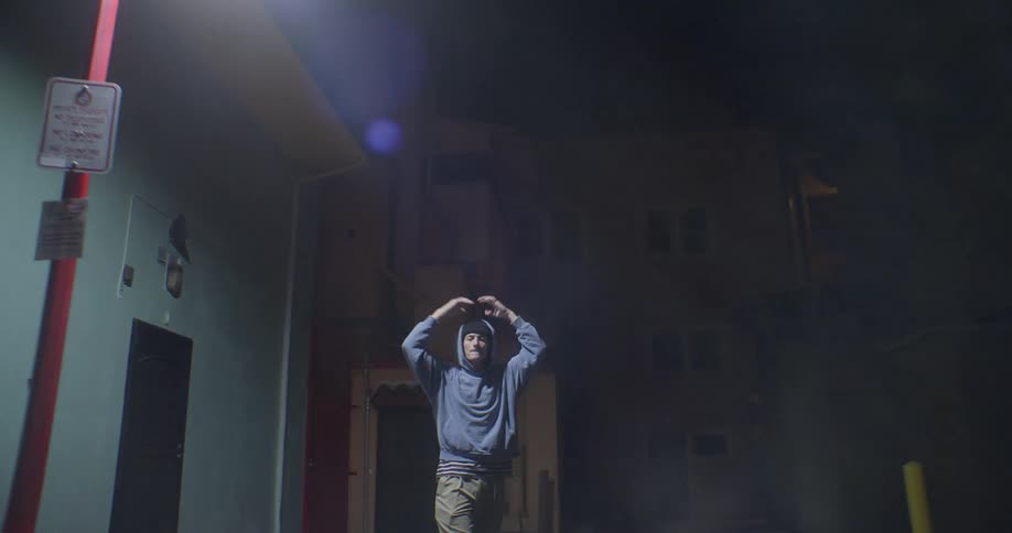 Man walking backward on the street at night with his hands raised pulls back his hood