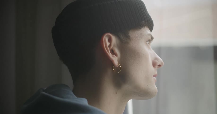 A young person wearing a black beanie and a gold hoop earring looks out a window with a thoughtful expression