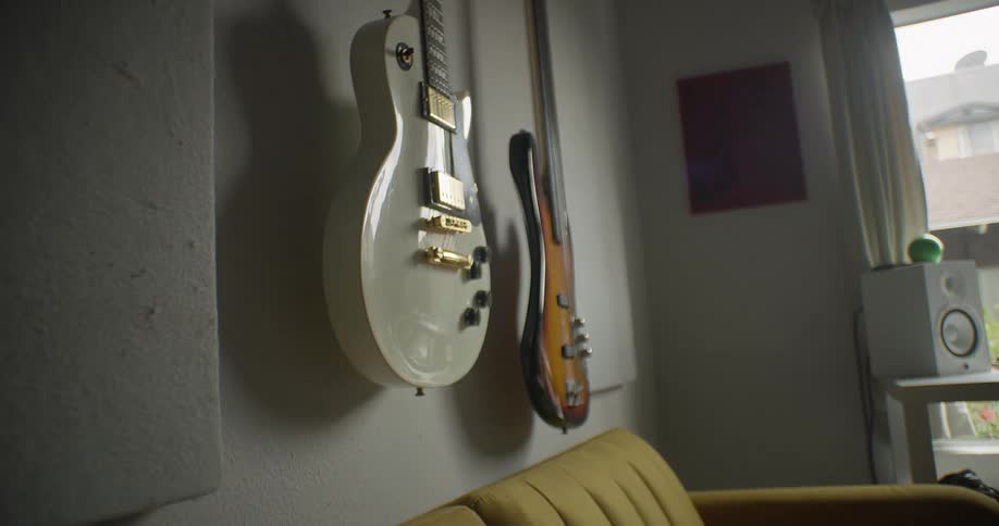 Electric and bass guitars hang on a wall above a yellow couch in a room with acoustic panels and window light