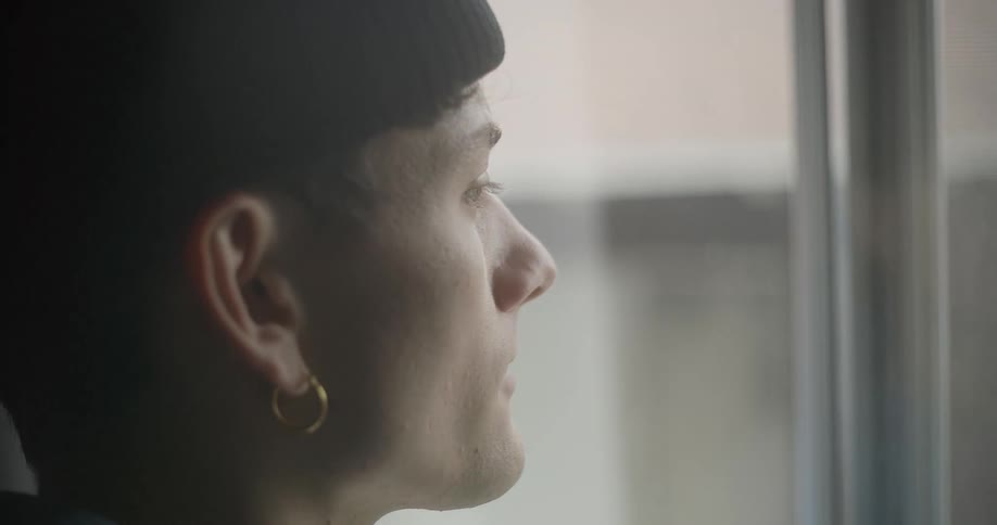 A young man with short hair wearing a black beanie and a gold hoop earring looking towards a window