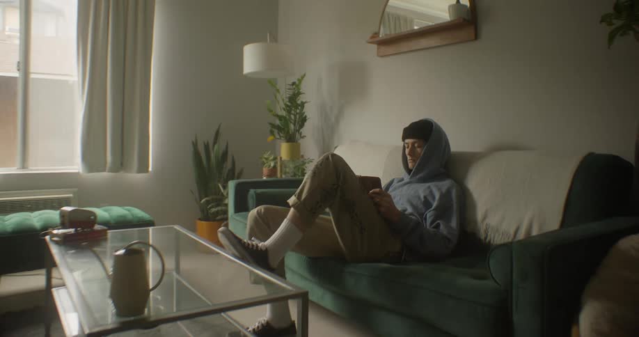 A young person wearing a black beanie and a light blue hooded sweatshirt sits on a couch in a softly lit room