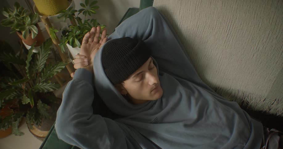 Young man lies on a couch located near a plant filled corner of the living room
