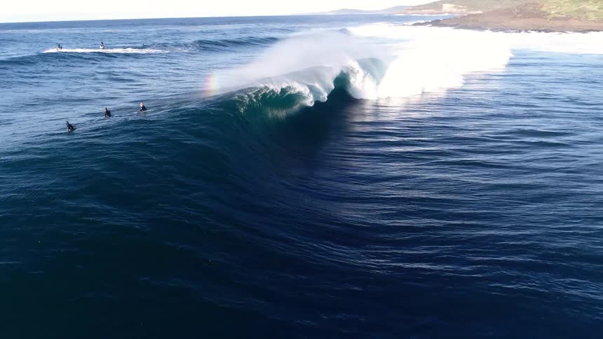 An aerial view captures several surfers paddling near a massive cresting ocean wave as it breaks creating spray with a visible rainbow while a jet ski waits nearby