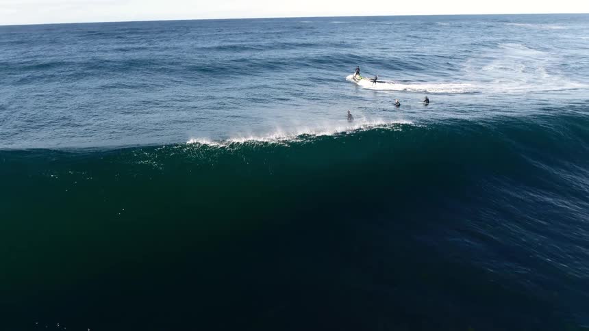 Several surfers navigate a large dark ocean wave as it breaks with one catching the ride near a jet ski