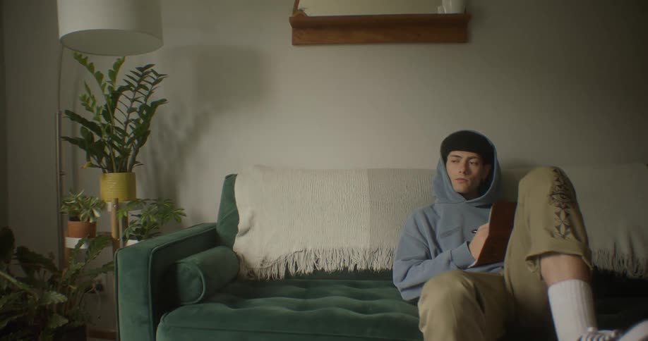 Young male sitting on a green couch he appears to be listening to music and writing in a journal the room has a few plants and the end table has an overhanging light there is a glass coffee table he is resting he foot on while he sits