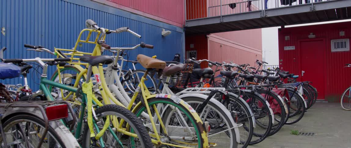 Two men walk past a group of bicycles parked next to a red and blue building