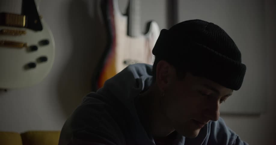 A young man sits on a sofa with two electric guitars on the wall behind him