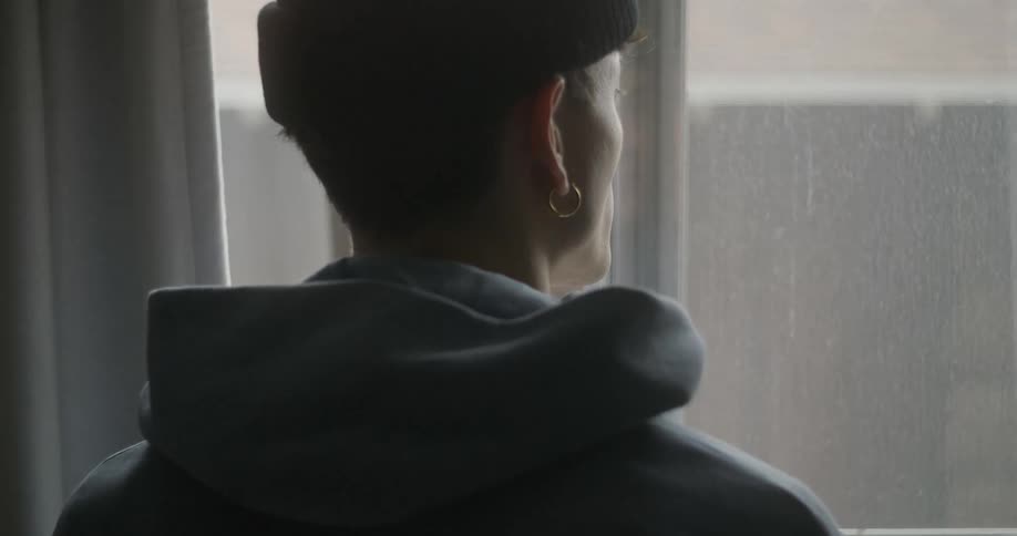 A young man stands inside an apartment and looks out the window