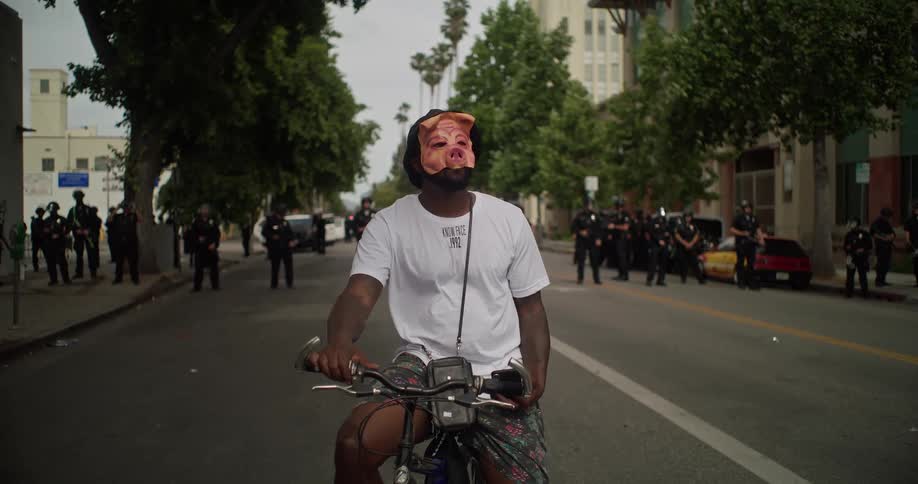 Man on a bicycle with a pig mask and in the background several police officers on a perimeter