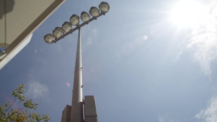 Looking up from a low angle a tall stadium light pole stands against a bright blue sky with the sun flaring beside it
