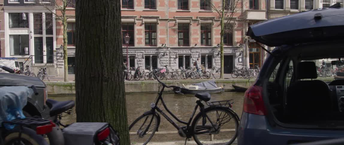 Two people stand near an open car across the river from several bicycles parked near a line of buildings during the daytime