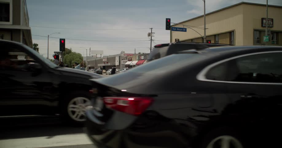 Traffic moves through a city as one car has a lot of smoke coming from it and a crowd on the corner watches
