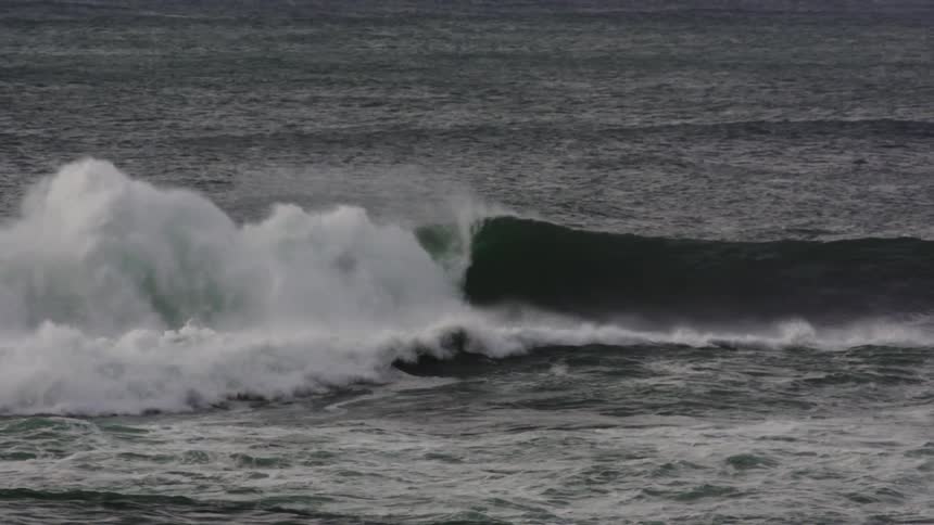 A large dark ocean wave curls and breaks in slow motion sending white spray into the air over the choppy sea surface