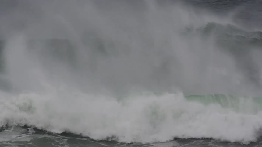 A large dark ocean wave crashes forcefully in slow motion throwing up white spray against the grey sea