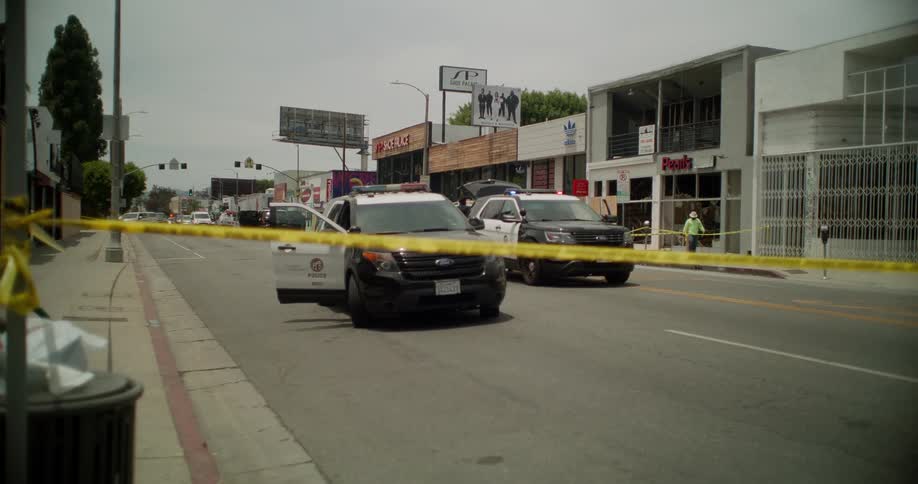 Cop cars sit in the street next to where a man crouches on the sidewalk in front of a business during the daytime