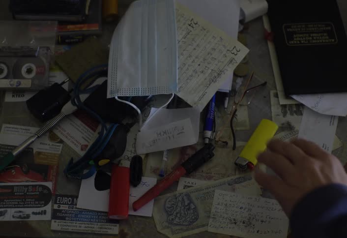 A hand picking up a yellow lighter from a desk scattered with items such as a face mask handwritten notes pens a lighter a cassette tape business cards and a passport