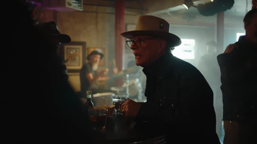 An older man in a hat talks intently with a woman at a table with drinks in a busy bar while a live band plays and people dance and watch in the background