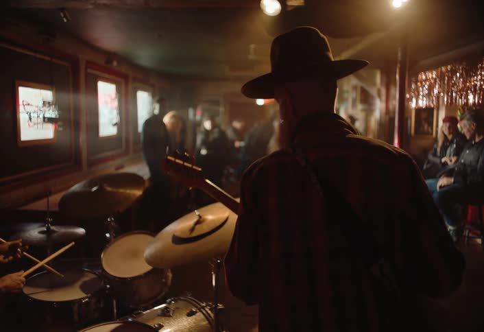 From behind a guitarist plays on stage next to a drumming bandmate in a dimly lit bar while audience members dance and talk