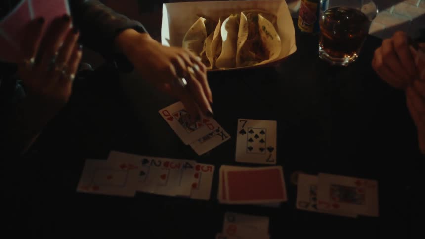 Friends play cards at a table holding tacos and a glass of liquor